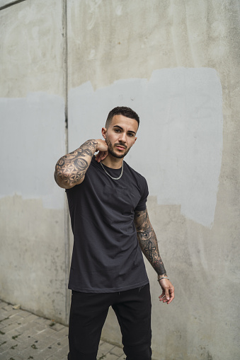 A vertical shot of a stylish European man with tattooed arms posing against a concrete wall