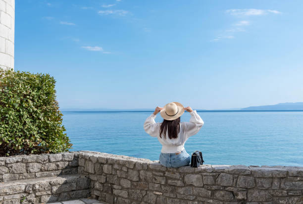 rear view of young woman sitting on stone wall on shore of sea. - beach stone wall one person imagens e fotografias de stock