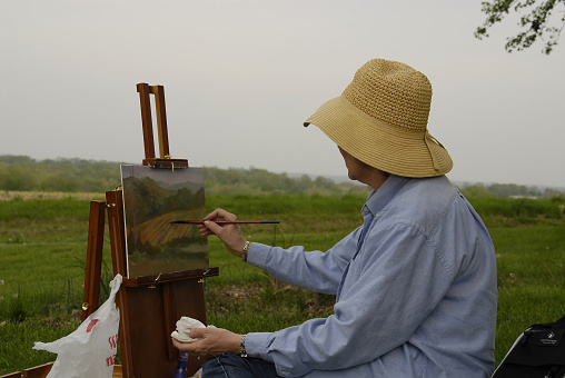60-year-old Latin woman with short white hair is sitting on a chair in the backyard of her house wearing a yellow apron doing her favorite hobby painting a beautiful picture on canvas