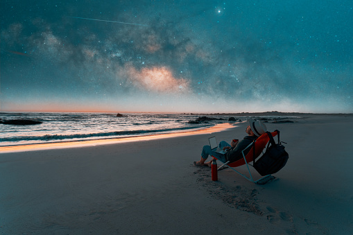 mature latin person sitting in a chair on the beach with a laptop drinking coffee and watching the stars in the Atacama sky