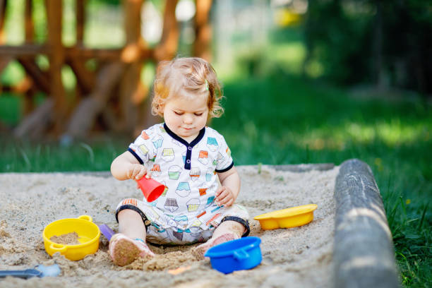 Cute toddler girl playing in sand on outdoor playground. Beautiful baby in summer clothes having fun on sunny warm summer day. Outdoors activity for toddlers. stock photo
