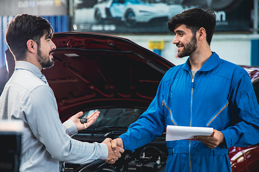 Man picking up car from garage a handsome male mechanic in a uniform returns the keys to the customer and shake hands with customers to thank.