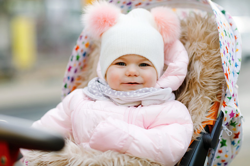 Cute little beautiful baby girl sitting in the pram or stroller on cold autumn, winter or spring day. Happy smiling child in warm clothes, fashion stylish baby coat and hat. Snow falling down.