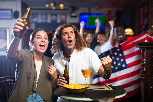 Expressive young adult couple sitting at table in sports bar, drinking beer and emotionally cheering for favorite team while watching american football match with fans waving USA flag in background