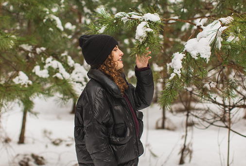 Redhead curly woman in black jacket and hat in winter pine forest. Pretty woman enjoying vacation. Girl laughing in nature. Feel happiness. Hoarfrost and snow on trees. Charming smile