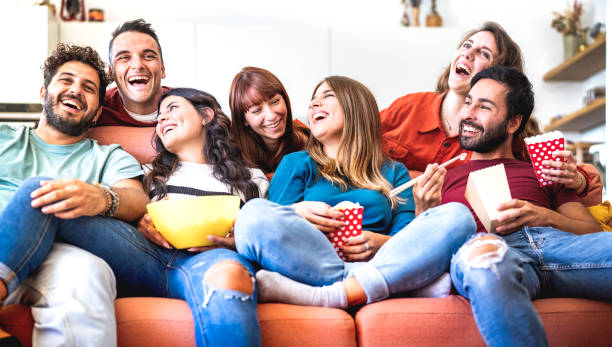 Young friends having genuine fun together at home watching movie on smart tv - Cozy life style concept with happy people relaxing on sofa couch eating popcorn and takeaway food - Bright vivid filter stock photo