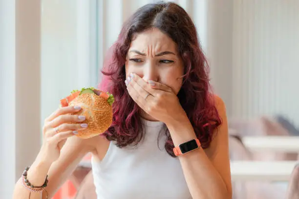 A girl eats a burger in a fast food cafe and feels nausea and heartburn. The concept of unhealthy food and food poisoning