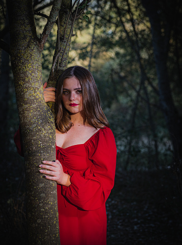 Portrait of an attractive young woman in a red dress, looking at the camera from behind a tree trunk in the forest.