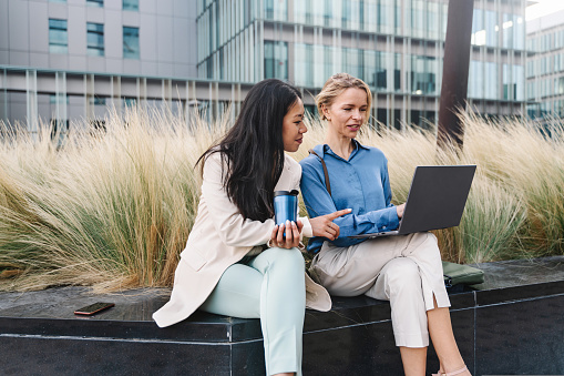 Two business women coworkers discussing project on laptop sitting together on bench outdoors in corporate office district