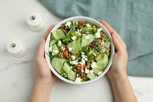 Woman holding bowl of delicious salad with lentils, vegetables and feta cheese at white table, top view
