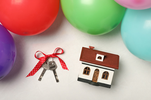 Wooden house model, keys with bow and balloons on white table, flat lay. Housewarming party