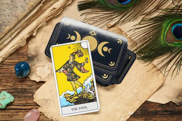 The Fool near other tarot cards with peacock feathers, gemstones and old book on wooden table, above view