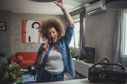 One woman, a cute middle-aged woman with curly hair is having fun at home singing karaoke, holding a microphone and listening to music