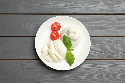 Delicious burrata cheese with basil and cut tomato on grey wooden table, top view