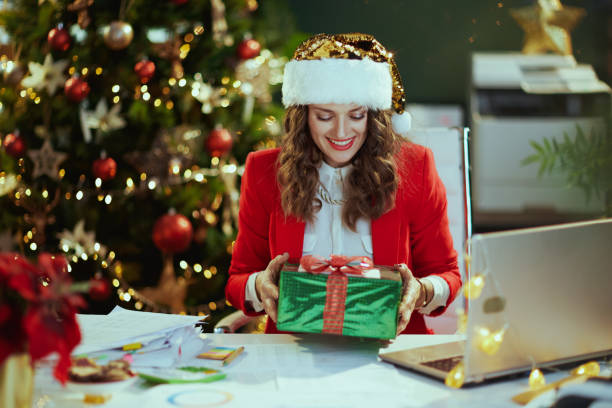 happy business woman in green office with Christmas tree Christmas time. happy modern 40 years old business woman in santa hat and red jacket with present box in modern green office with Christmas tree. perfect gift stock pictures, royalty-free photos & images