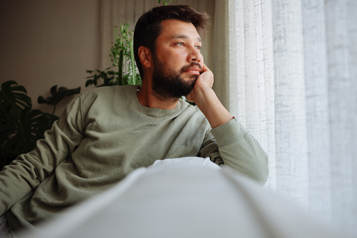 Man in living room looking through window, being thoughtful