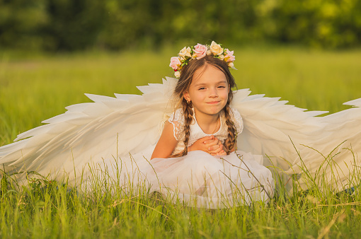Angel with white wings on green grass. Blonde girl in dress on summer background