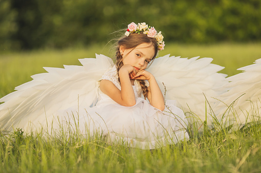 Angel with white wings on green grass. Blonde girl in dress on summer background