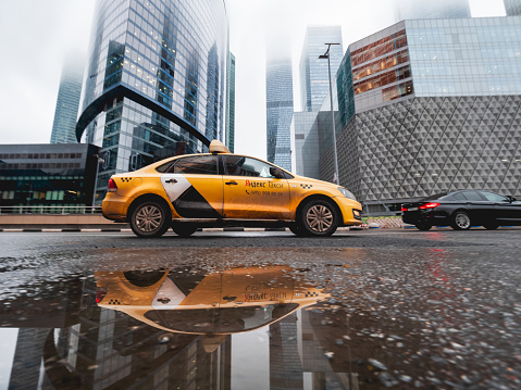 Moscow, Russia - November 09, 2022. Yandex Taxi is moving on urban road among skyscrapers of Moscow International Business Center. Car's reflection in puddle on wet road bed. Rainy weather in town.