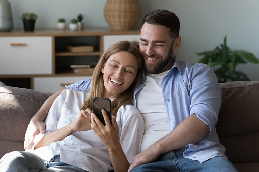 Beautiful young wife holds smartphone web surfing information spend carefree weekend with husband relax on couch at cozy home. Buying goods on-line, make remote order, enjoy retail e-services concept