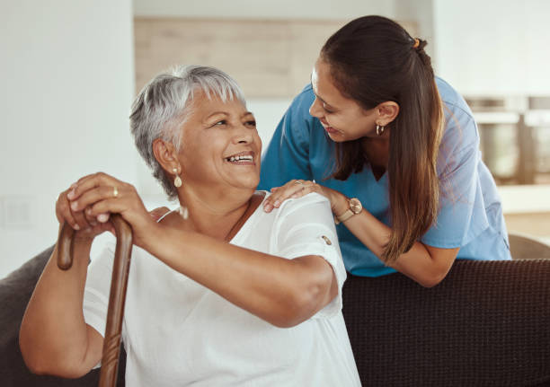 happy, relax and senior woman with caregiver smile while sitting on a living room sofa in a nursing home. support, help and professional nurse or healthcare worker helping elderly lady or patient - ouderenzorg stockfoto's en -beelden