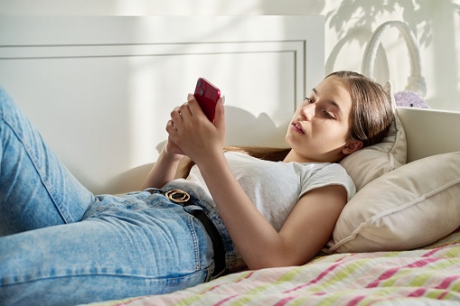 Teenage girl lying in bed using smartphone for leisure communication, adolescent teen female texting on phone at home in her bedroom. Lifestyle, gadget applications for leisure communication study