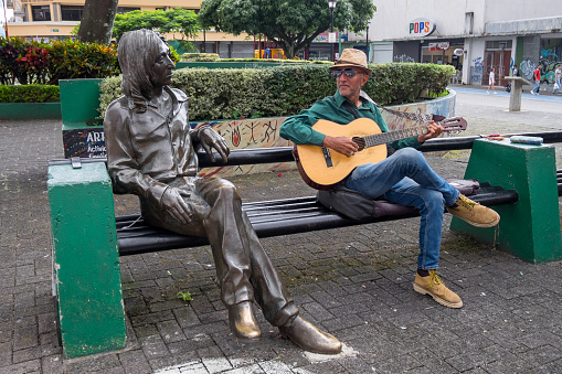 San José, Costa Rica - September 16, 2022: A man plays the guitar sitting next to a statue of John Lennon, created by Cuban sculptor José Ramón Villa, and placed in the Plaza de las Artes in Chinatown, in the urban center of the city