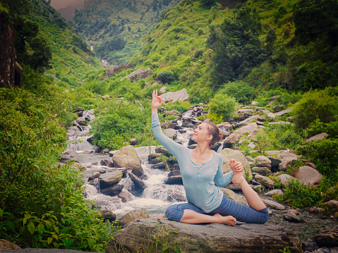 Yoga outdoors - young sporty fit woman doing stretching yoga asana Eka pada rajakapotasana - one-legged king pigeon pose at tropical waterfall. Vintage retro effect filtered hipster style image.