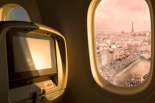 Paris City, France at sunset / sunrise sky aerial view from porthole window airplane economic seat after take off from airport. On back, the Eiffel Tower, Arch de Triomphe in Paris Downtown District, Travel concept. Plane interior.