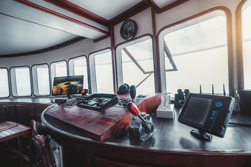 A wide-angle perspective view of a vessel dashboard with plenty of instruments, dial indicators, and other navigational equipment; a yacht control panel on the captain's bridge with a compass