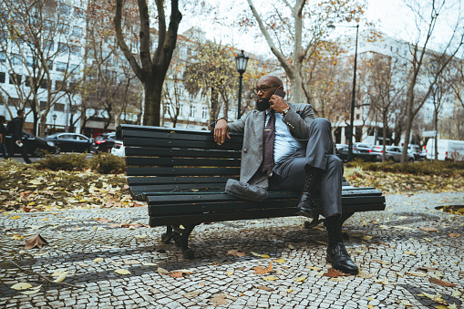 A wide-angle shot with a handsome mature bald African American man entrepreneur with a black beard, having a business conversation via his smartphone while sitting on a park bench on an autumn day