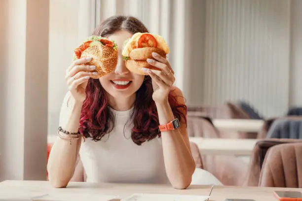 A girl in a cafe fooling around and closed her eyes with two cheeseburgers. Fast food and healthy diet concept