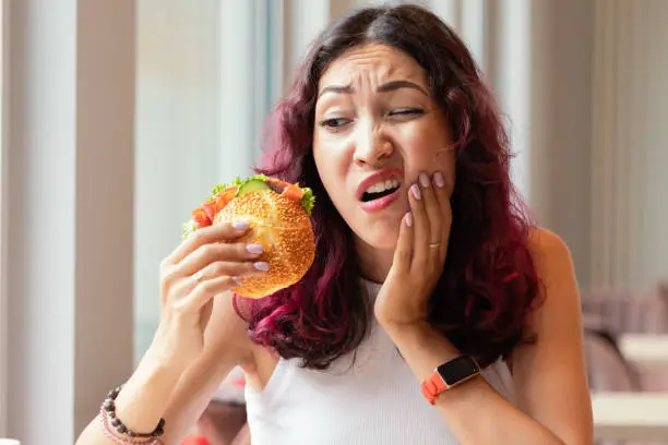 Woman eating a hamburger in a fast food cafe and felt a sharp pain in her mouth - she bit her tongue or cheek, stomatitis and dental problems or jaw joint disease