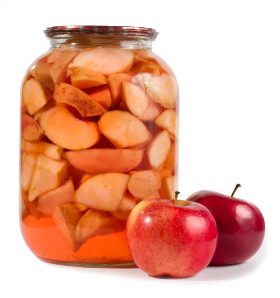 Canned apples in a glass jar on a white background. Fresh apples are in the foreground.