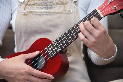 Young woman playing red ukulele at home, sitting on the floor, dressed as a hipster, learning songs.