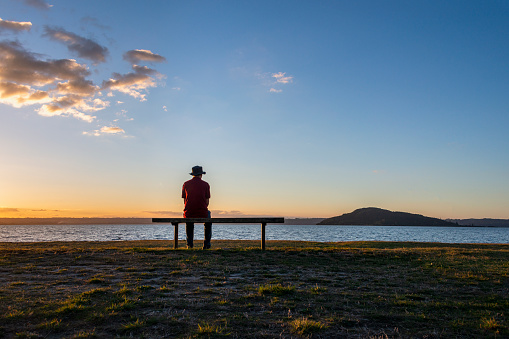 Man sitting alone by lake Rotorua at sunset, Mokoia Island in the distance, Rotorua.