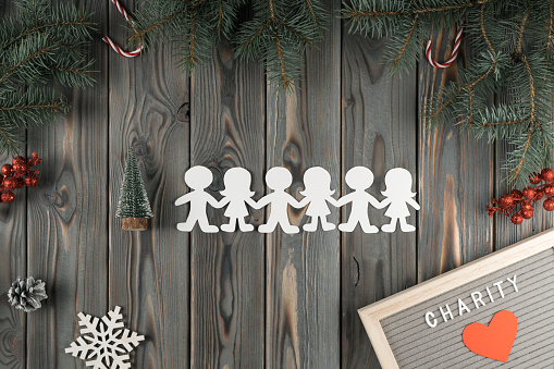 Group of paper cut people on gray wooden wall, top view. Helping needy concept. Decor from fir branches with snowflakes, cones and lollipops. Giving money. New Year or Christmas charity, kind heart.