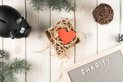 Black piggy bank for donation and present box with red paper heart postcard, top view. Decorative spruce branches on white planking. Giving money. Kindness and Christmas or New Year charity concept.