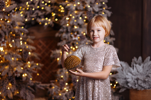 A girl in a silver glittery dress decorates the Christmas tree in a cozy living room of a country house with a stone fireplace.