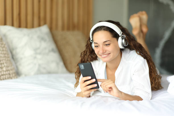 donna che usa il telefono che indossa le cuffie sul letto - hotel wireless technology bedroom hotel room foto e immagini stock
