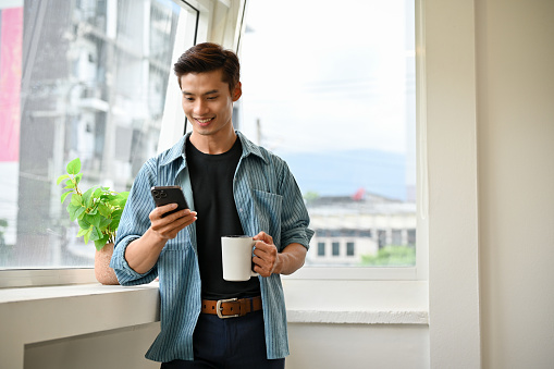 Handsome millennial Asian male boss or office employee in casual clothes is on his coffee break, sipping coffee and using smartphone while standing by the window.