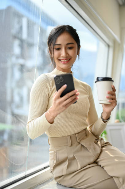 istock Attractive Asian female sitting on window bench, enjoying her coffee and using smartphone. 1441348019