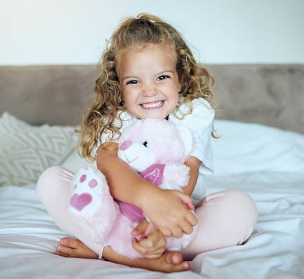 Children, teddy bear and girl with a child hug her stuffed animal with a smile in her house. Portrait of kid, happy and safe with an adorable or cute female holding a fluffy toy sitting on a bed