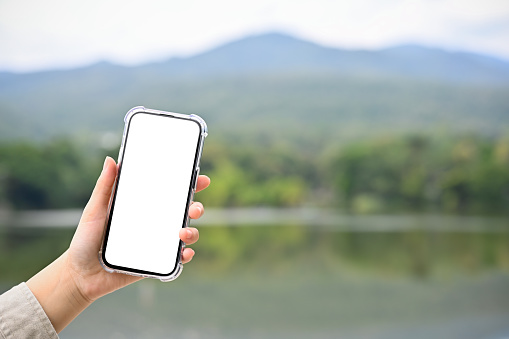A smartphone white screen mockup is in a woman's hand over blurred background of beautiful green mountains and lake. close-up image