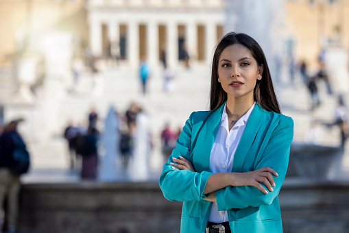 A young, confident business woman with crossed arms stands in front of a public building in a city