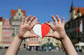 A red-white heart in the colors of the national flag of Poland in the hands of a child against the backdrop of the old town square. Independence Day of Poland. Freedom and Democracy