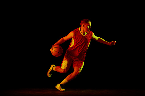 Dynamic portrait of young active athlete, male basketball player in sports uniform practicing with ball, jumping, dribbling isolated over dark background in neon light. Copy space for ad