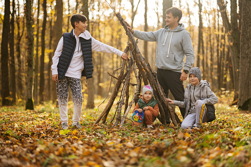 Children with father constructs a house from sticks in autumn forest.