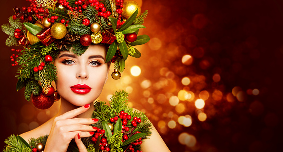 Christmas Woman Beauty. Beautiful Model in Fir Tree Wreath with Xmas Ornaments. Women Face Skin and Hands Winter Care. Fashion Girl with Red Lips Make up over Shining Lights Background