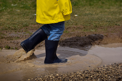 A photo showing a child's legs playing with a puddle outdoors on a rainy day. The child is wearing blue rain boots.